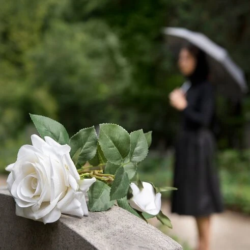Weiße Rosen als stilles Zeichen der Erinnerung auf einem Grabstein, im Hintergrund eine trauernde Person mit Regenschirm vor grüner Natur
